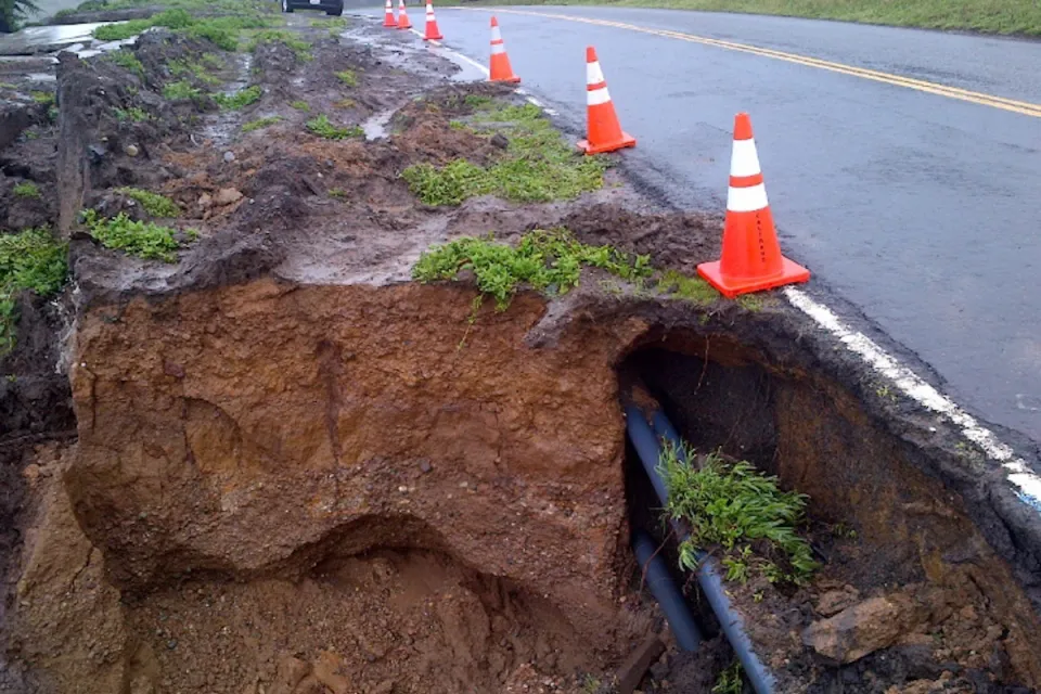 Eroded cliffside and exposed utilities along asphalt Highway 1 in California with orange traffic cones lining side of road.